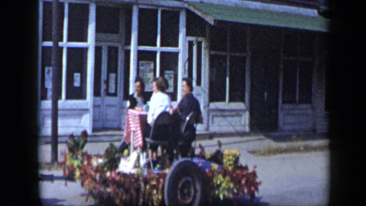 Women in period clothing ride or stand on a decorated parade float in Aledo, Texas, with storefront windows behind them.