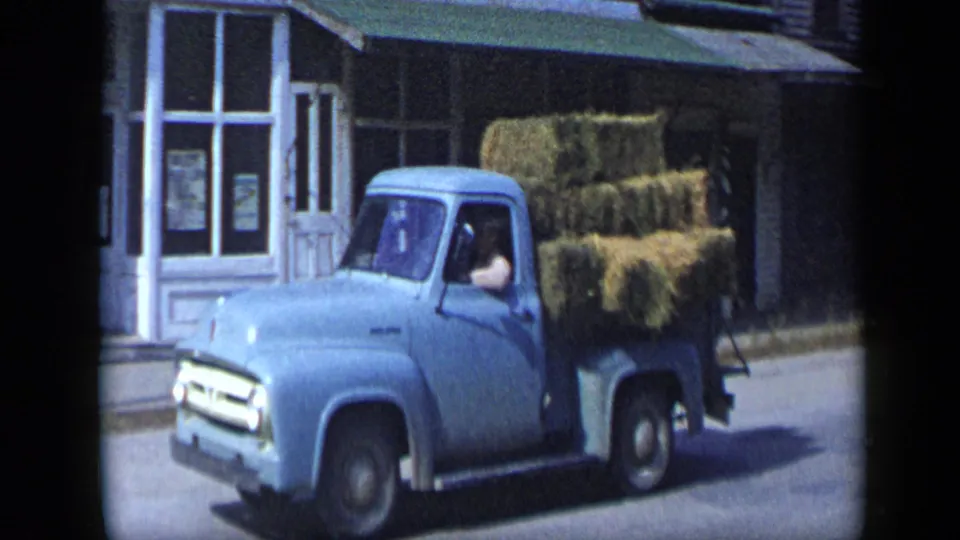 A blue pickup carrying stacked hay bales moves along an Aledo, Texas street, with the driver visible through the cab window.