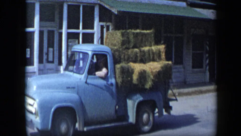 The blue hay truck continues past storefront windows on the Aledo street, with the driver partly visible in the cab.