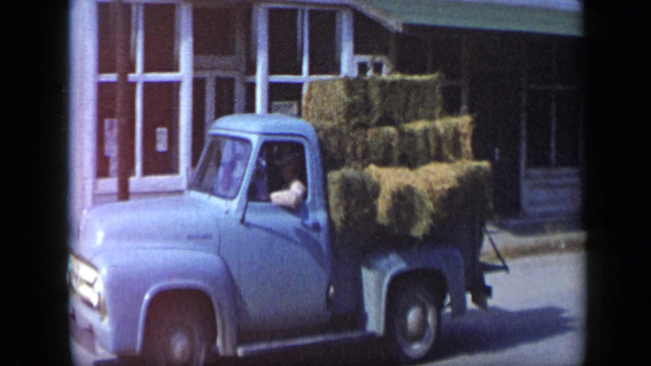 In 1959 Aledo, Texas, a light blue pickup carrying stacked hay bales passes a white storefront as the driver sits in the cab.
