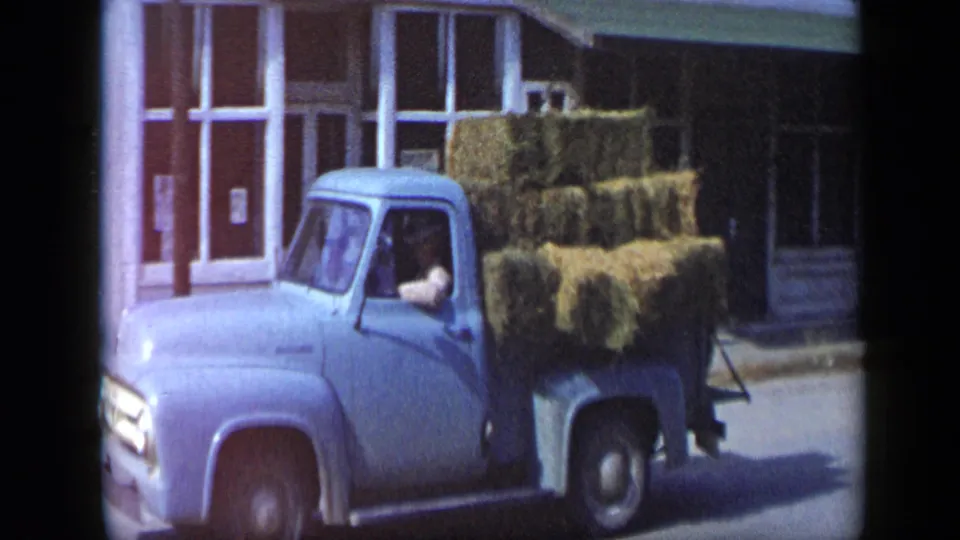In 1959 Aledo, Texas, a light blue pickup carrying stacked hay bales passes a white storefront as the driver sits in the cab.