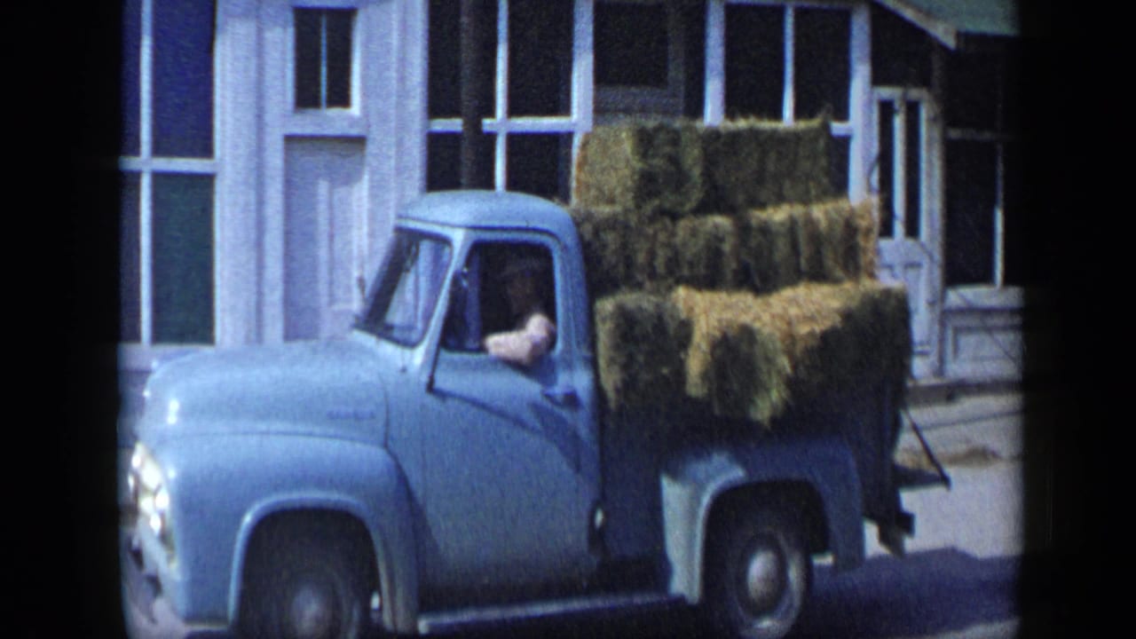 The blue pickup continues past the Aledo storefront with hay bales stacked high behind the cab and a driver visible through the window.
