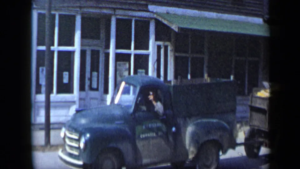 A darker pickup or farm truck tows a wooden trailer filled with yellow produce along the storefront-lined street in Aledo, Texas.