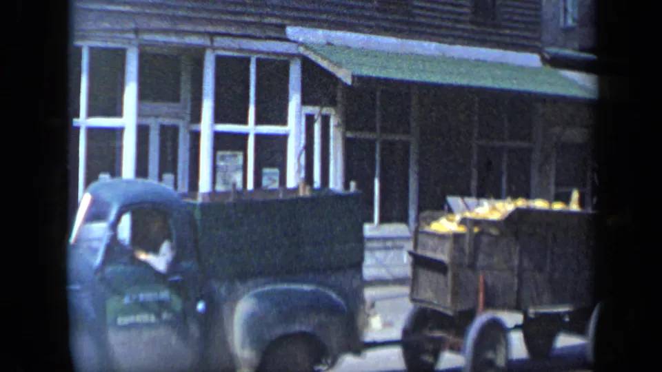 The vehicle is partly hidden at frame edge while a wooden trailer loaded with yellow produce passes the storefronts in the 1959 Aledo street scene.