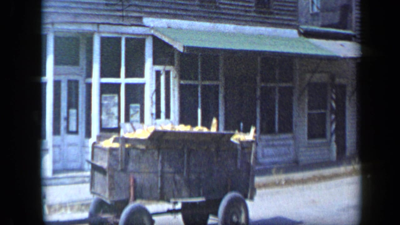Aledo, Texas 1959 frame with a wooden wagon or trailer holding yellow produce in front of a small storefront with a green awning.