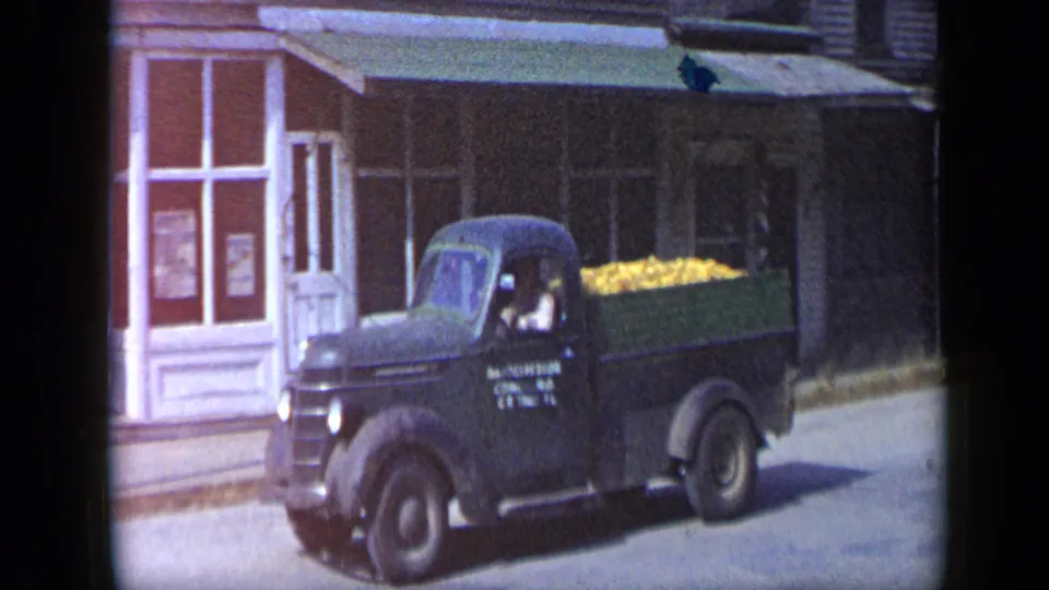 Aledo, Texas 1959 frame showing a black pickup loaded with yellow produce, a visible driver, and storefront windows behind it.