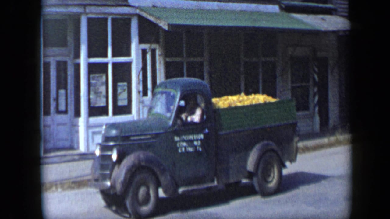Dark green pickup truck carrying yellow produce passes storefront windows.