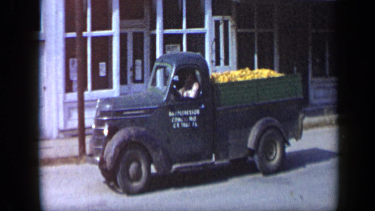 Green produce truck moves past storefronts with a driver visible.