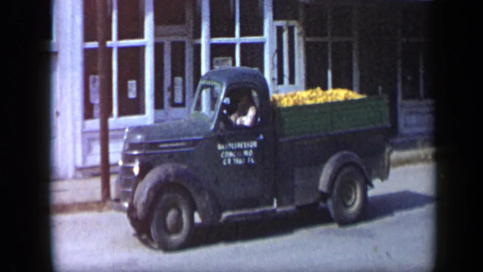 The green pickup loaded with yellow produce continues along an Aledo storefront street, with the driver visible through the cab window.