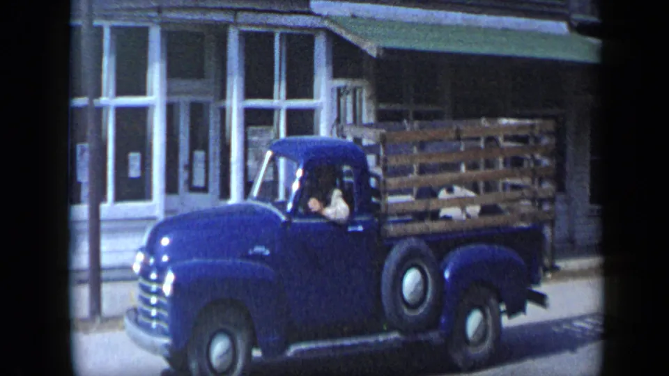A blue mid-century pickup with wooden side rails passes the storefront in the 1959 Aledo, Texas street scene.