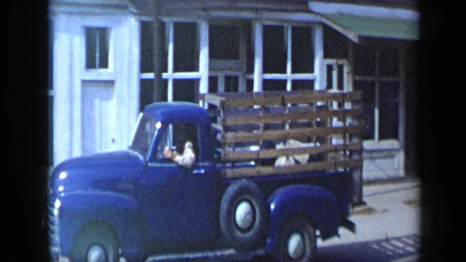 The blue pickup with wooden rails continues past storefront windows, with a driver visible inside the cab.