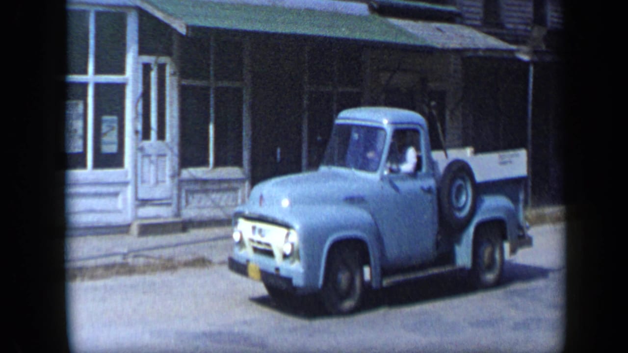 1959 Aledo, Texas frame with a light blue pickup and side-mounted spare tire passing storefront windows and awnings.