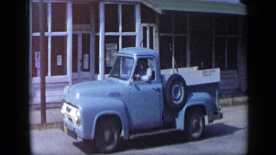 Late-1950s Aledo, Texas home-movie frame of a blue pickup continuing down a storefront-lined street.