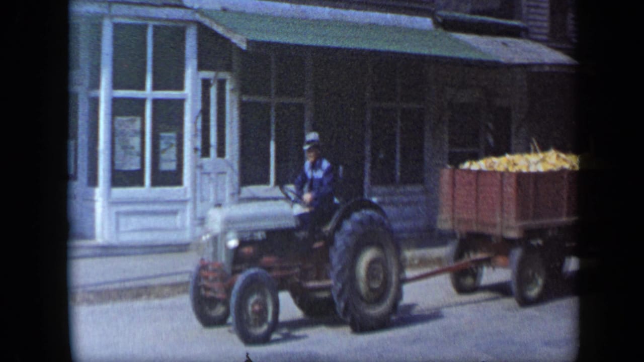 Man drives a red tractor pulling a corn-filled wooden wagon past a storefront.