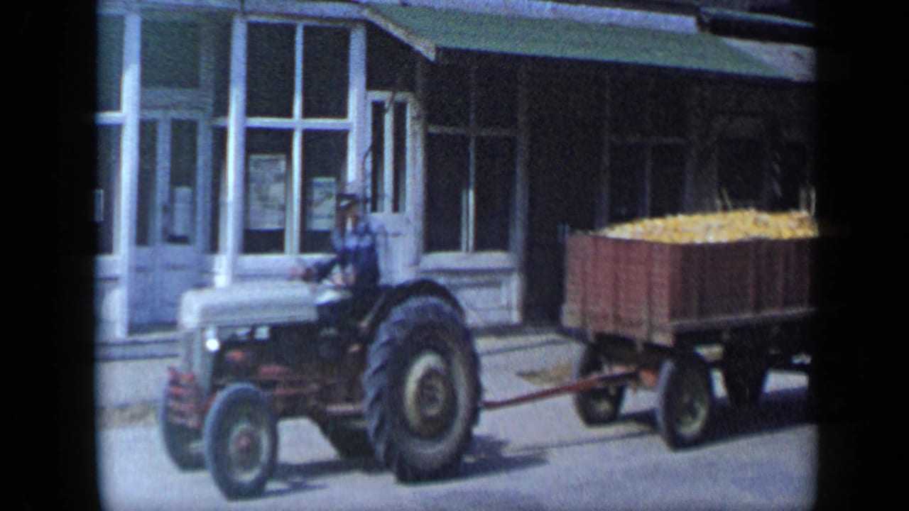 Tractor and corn wagon continue along the street beside a windowed building.