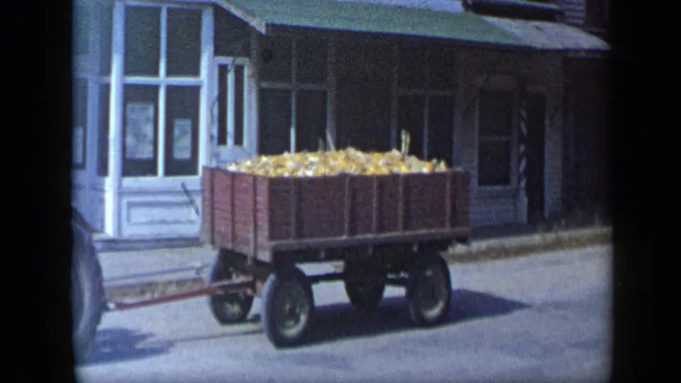 A wooden wagon piled with harvested corn moves through the frame behind the mostly off-screen tractor in the 1959 Aledo footage.