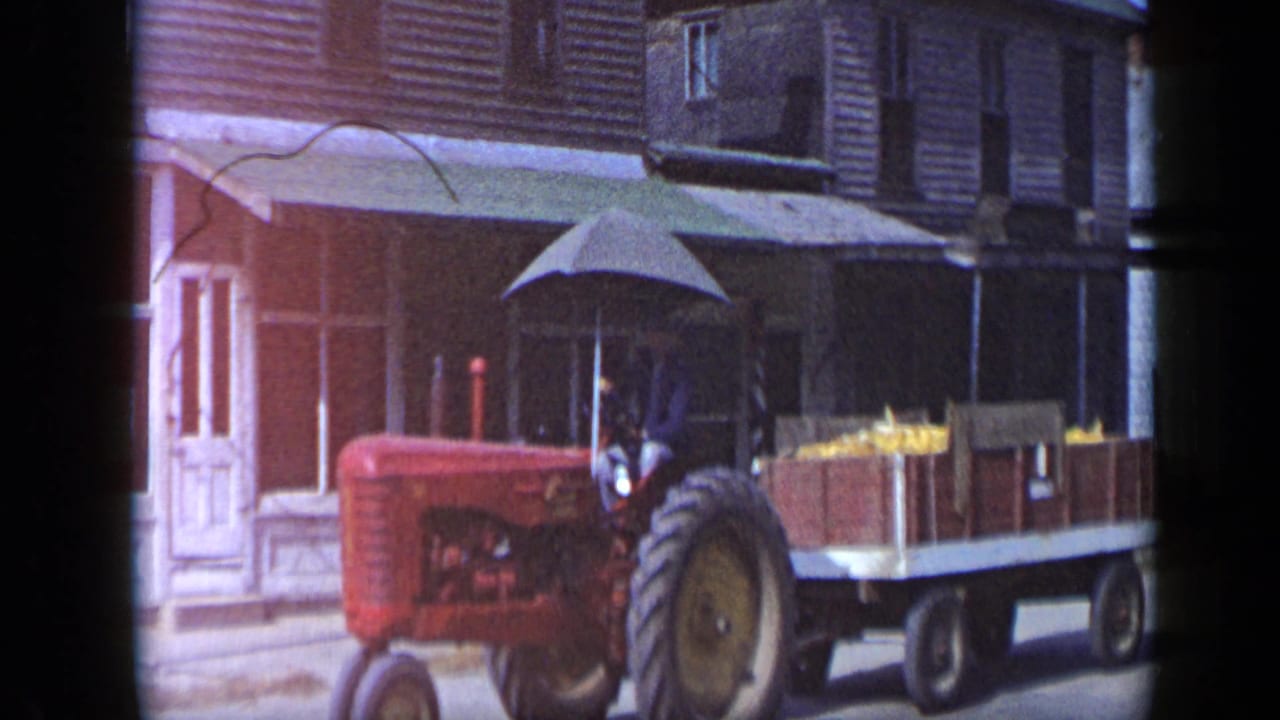 In Aledo, Texas, 1959, a red tractor with a driver pulls a wooden produce wagon along a small-town street.