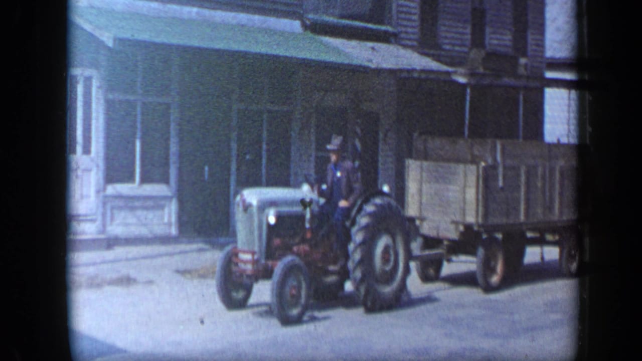 In Aledo, Texas in 1959, a man drives a vintage tractor pulling a wooden wagon along a quiet street.