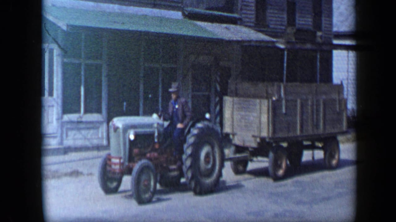 The tractor and wooden wagon continue along the Aledo street, with the driver seated beside storefront windows and a green awning.