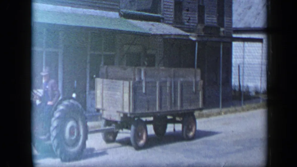 Aledo, Texas 1959 home-movie frame showing a tractor towing a wooden trailer past streetfront buildings.