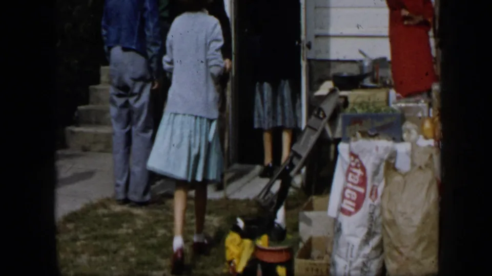Aledo, Texas 1959 home-movie frame showing adults gathered by steps beside supplies, sacks, jars, and produce outdoors.