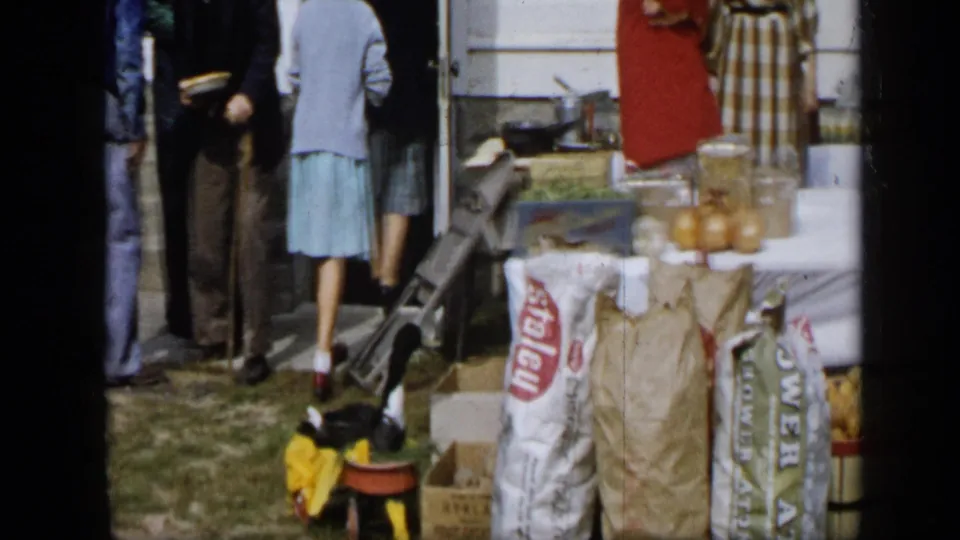 Aledo, Texas 1959 frame showing people in period clothing beside sacks, jars, boxes, and produce at an outdoor gathering.