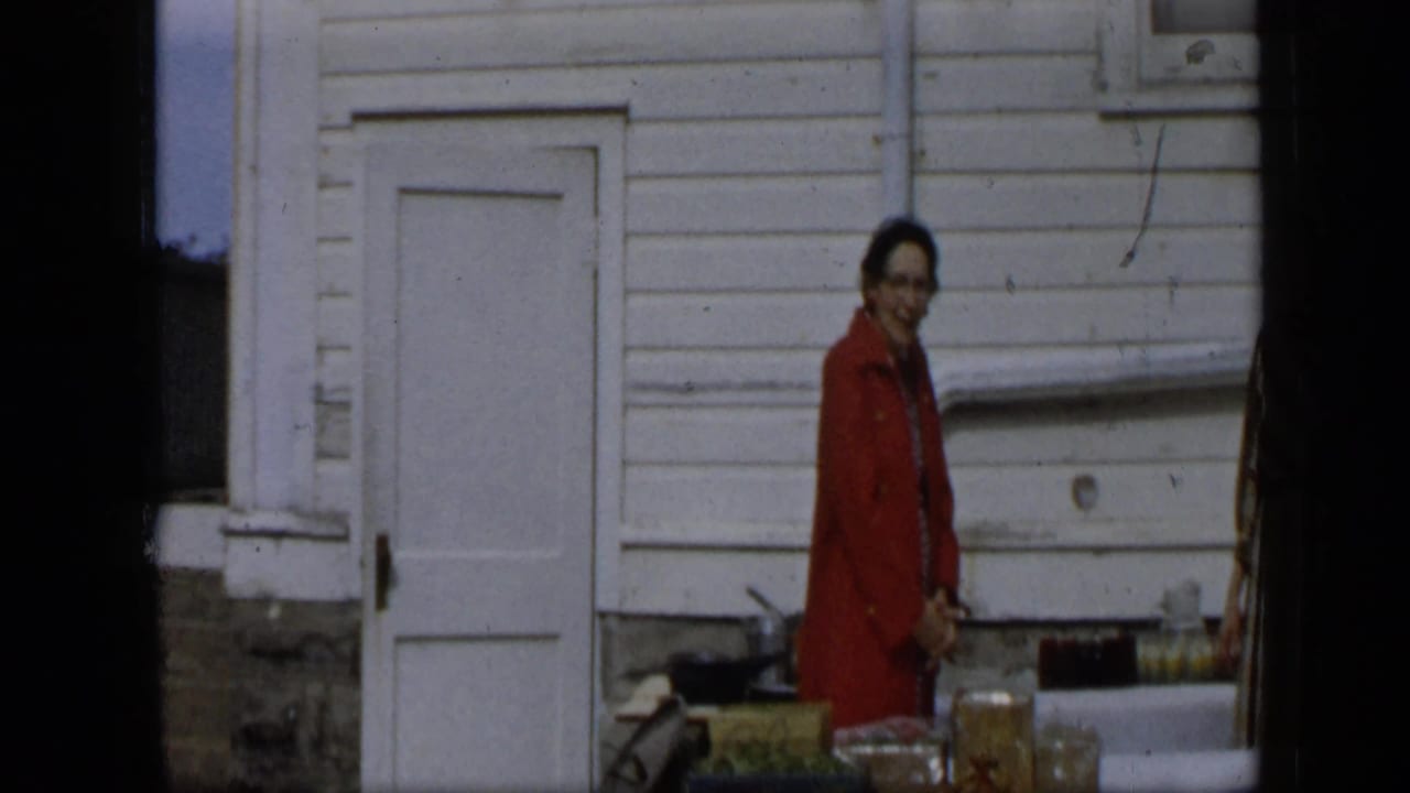 Woman in a red coat standing by an outdoor produce table against a white clapboard wall.