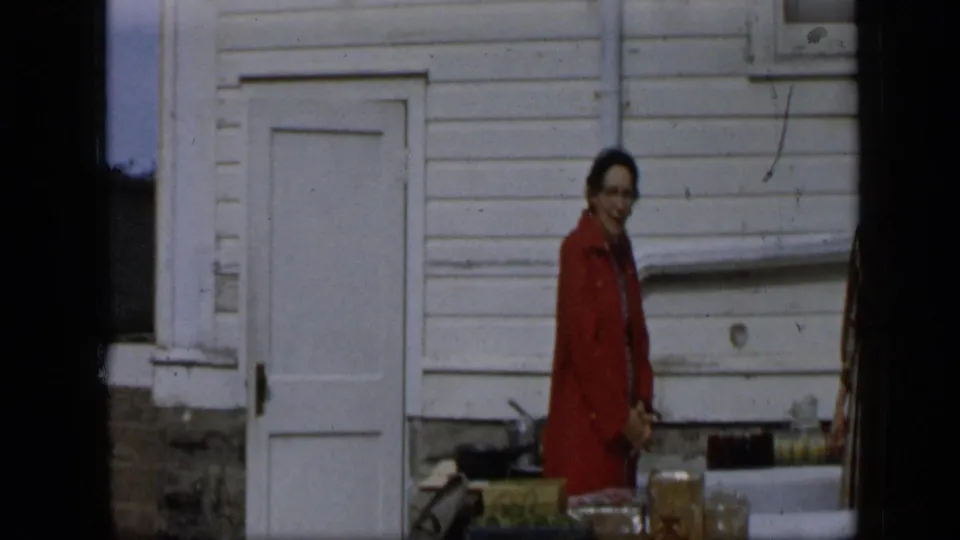 In the supplied 1959 Aledo, Texas context, a woman in a red coat stands beside boxes and produce outside a white clapboard building.