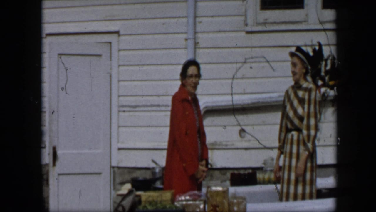 Two women stand outside beside a table, one in red and one in a checked dress with a hat.