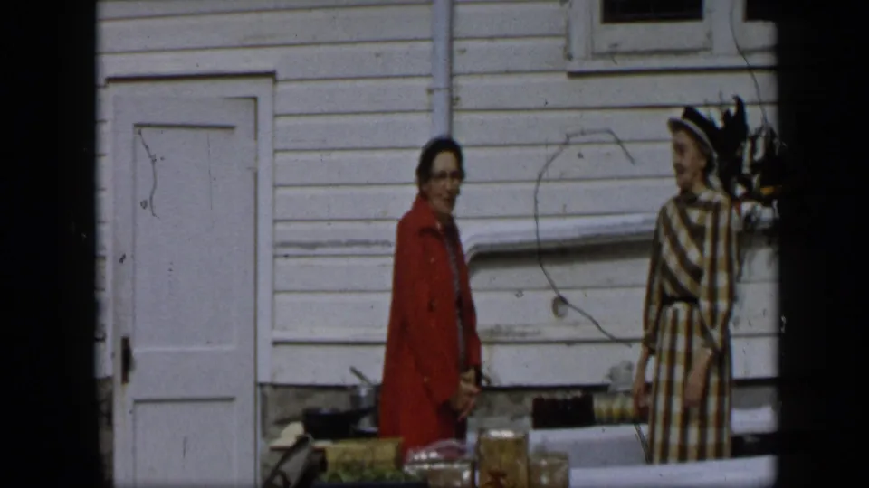 Two adult women stand at an outdoor produce setup in Aledo, Texas, one in a red coat and one in a checked dress and hat.
