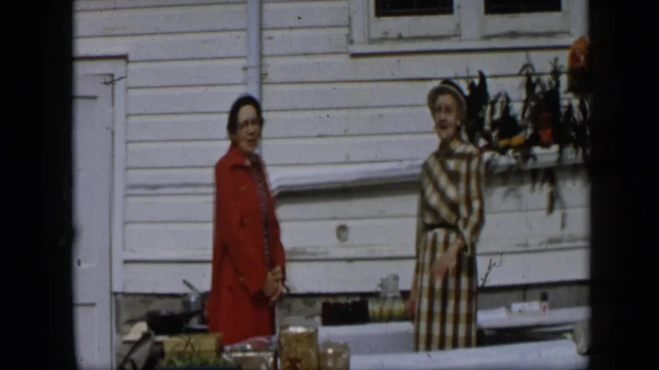 The two women remain beside the produce table, with boxes, containers, siding, and a decorative hanging visible behind them.
