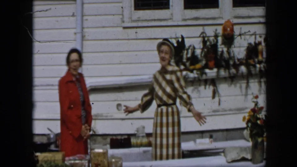 A woman in a checked dress and hat gestures with open arms beside a woman in red at the 1959 Aledo outdoor produce scene.