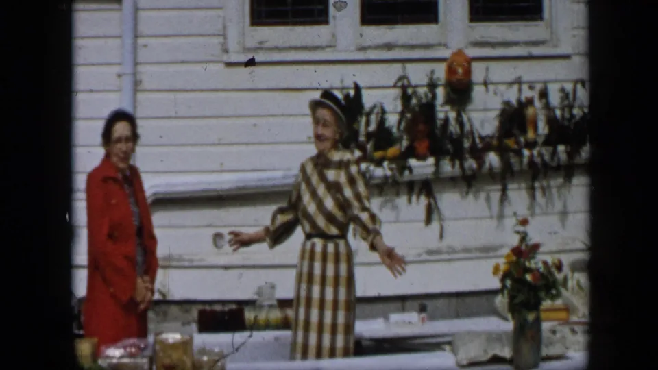 In Aledo, Texas, 1959, two women stand near a display table outside a white clapboard house with jars, flowers, and fall decor.
