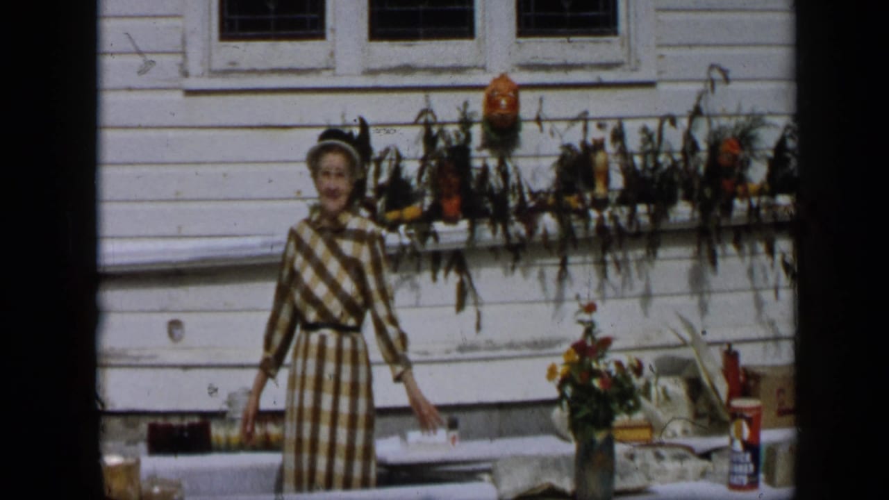 In Aledo, Texas, 1959, a woman in a brown and white checked dress gestures beside an outdoor table beneath autumn window decor.