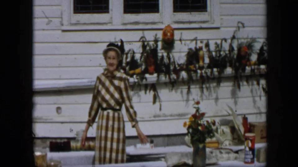 In Aledo, Texas, 1959, a woman in a brown and white checked dress gestures beside an outdoor table beneath autumn window decor.