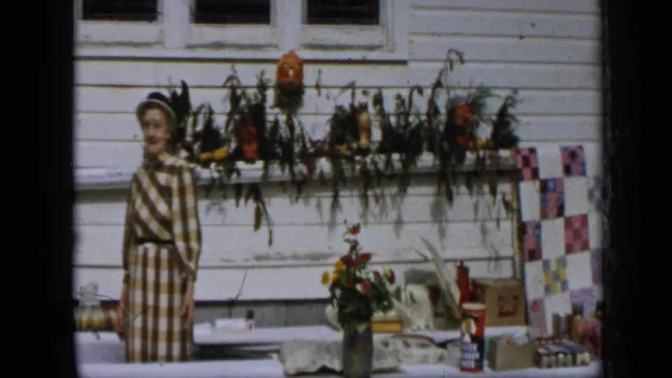 In Aledo, Texas, 1959, a woman stands by an outdoor sale table while a colorful quilt or textile appears along the wall nearby.