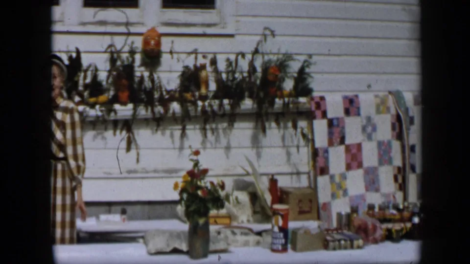 In Aledo, Texas, 1959, the frame shows a decorated outdoor table, a quilt or textile, and a partly visible woman near a white clapboard house.