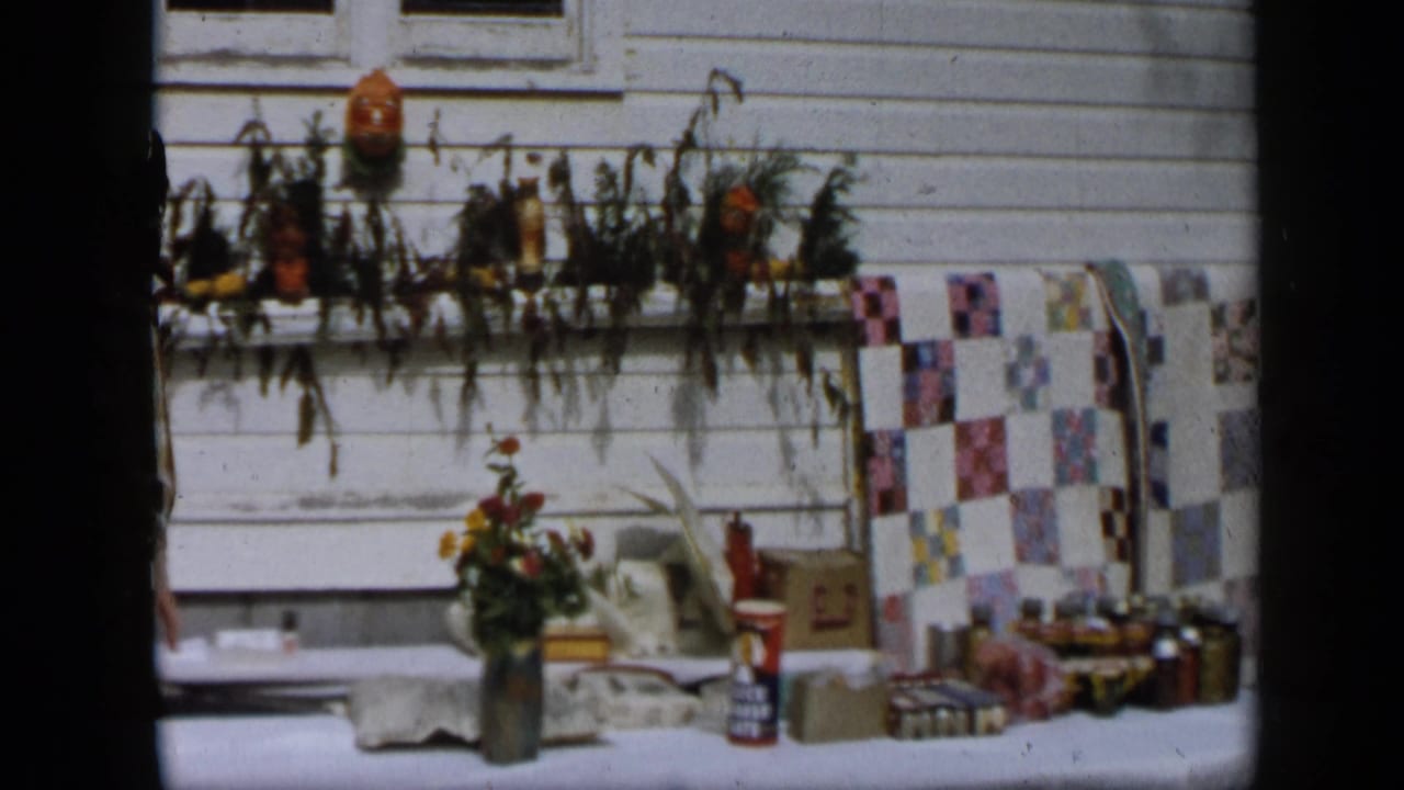 Aledo, Texas 1959 home-movie frame showing a porch display with flowers, boxes, patterned fabric, and household items.