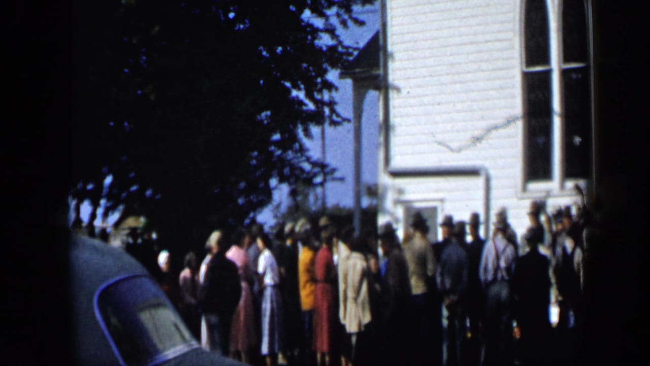 Crowd in mid-century clothing waits outside a white clapboard building in Aledo, Texas, in this 1959 home-movie frame.