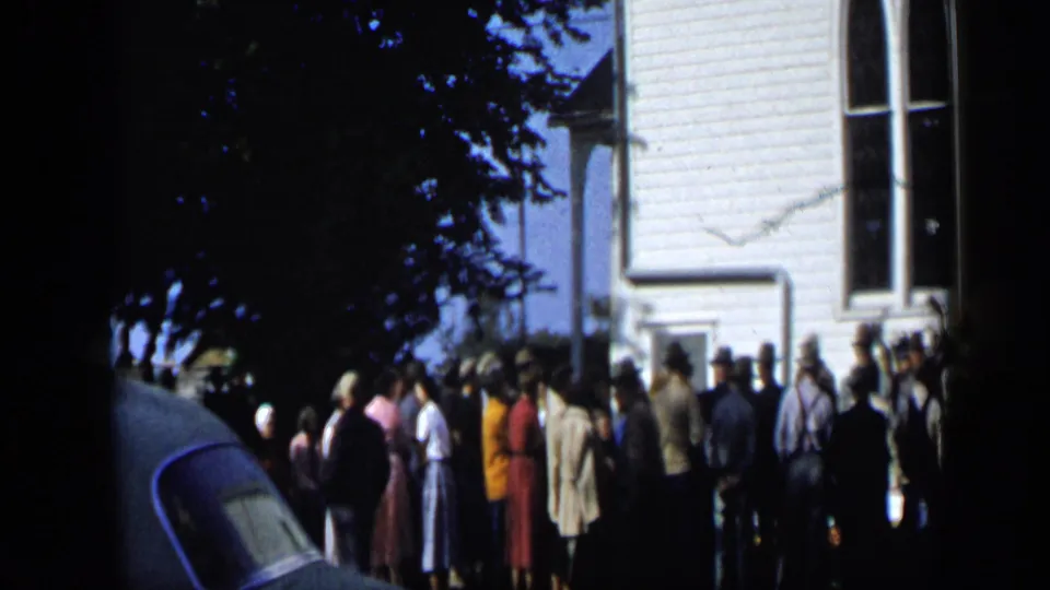 Crowd in mid-century clothing waits outside a white clapboard building in Aledo, Texas, in this 1959 home-movie frame.
