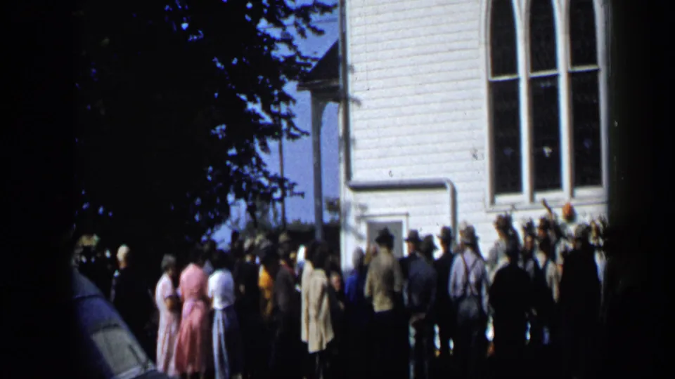 1959 Aledo, Texas frame of a crowd waiting beside a white clapboard building, with trees shading part of the scene.