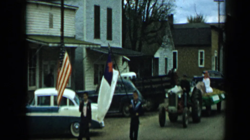 Aledo, Texas, 1959 parade view with a tractor pulling a hay trailer past parked cars and roadside flags.