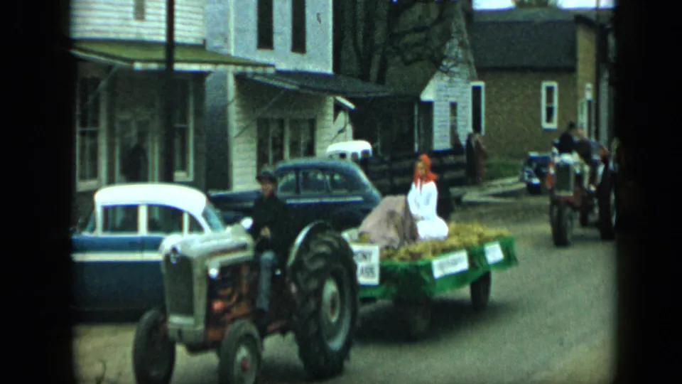 Aledo, Texas, 1959 frame showing a tractor and hay-filled trailer with riders moving along a residential downtown street.