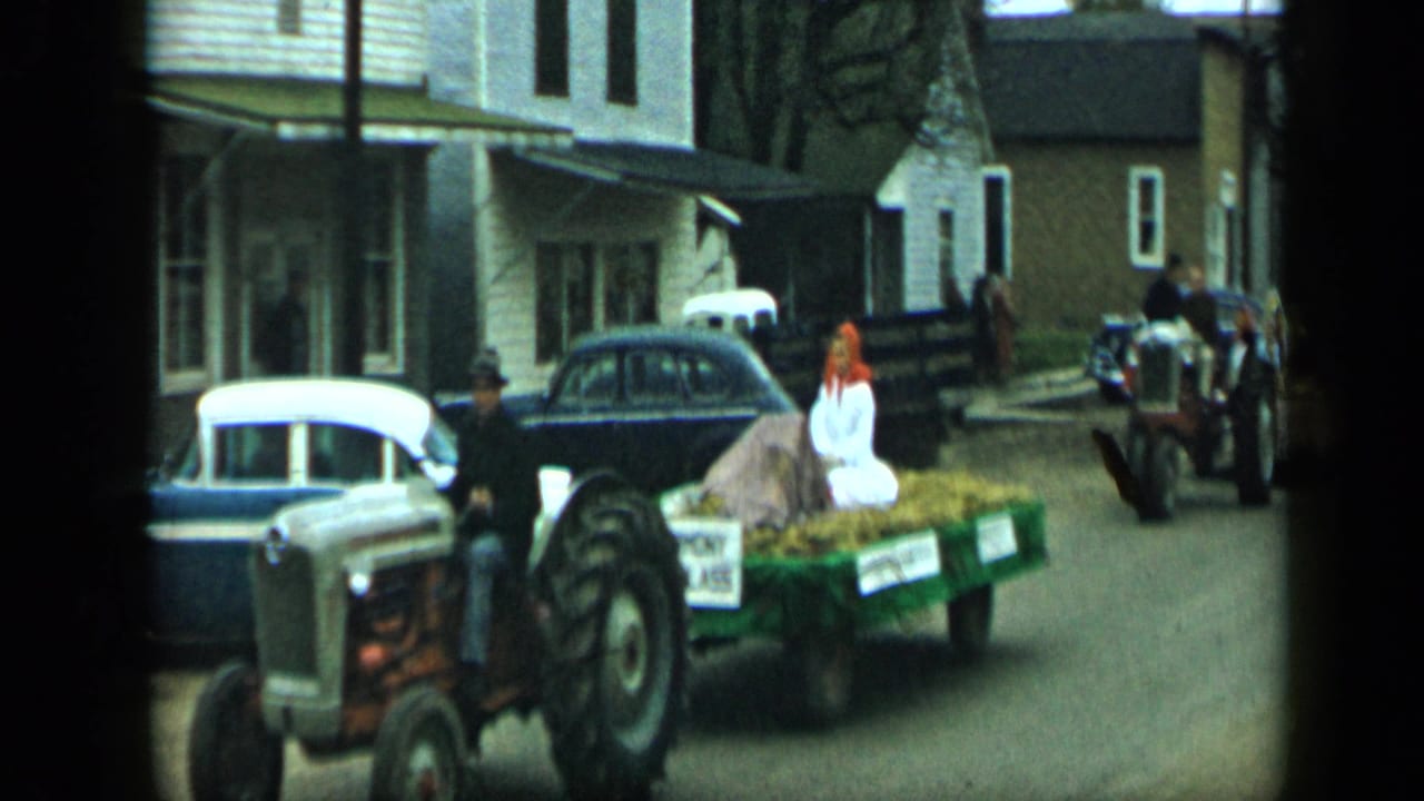 In Aledo, Texas in 1959, a tractor pulls a green hay float with a seated woman past classic cars and houses.
