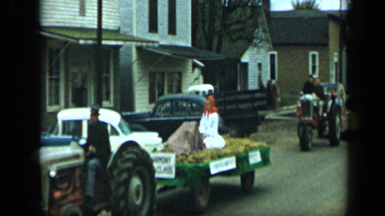 A 1959 Aledo parade float carrying a woman in white moves along a residential street with tractors and mid-century cars nearby.