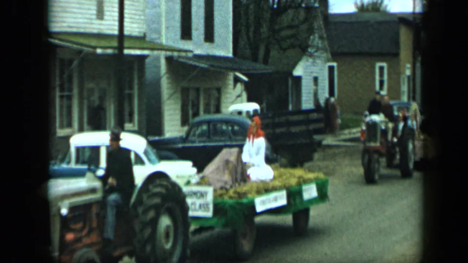 A 1959 Aledo parade float carrying a woman in white moves along a residential street with tractors and mid-century cars nearby.