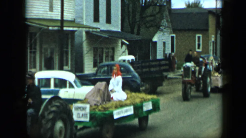 The Aledo, Texas parade float moves closer to camera, with a woman seated on hay and a tractor partly entering the frame.