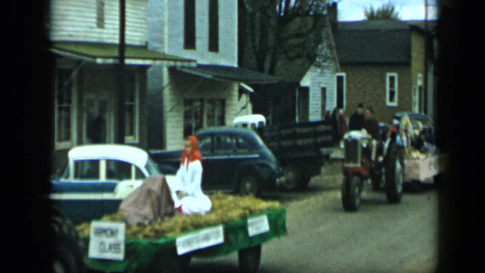 A tractor-pulled float in the 1959 Aledo, Texas parade passes classic cars while another tractor and riders follow in the background.