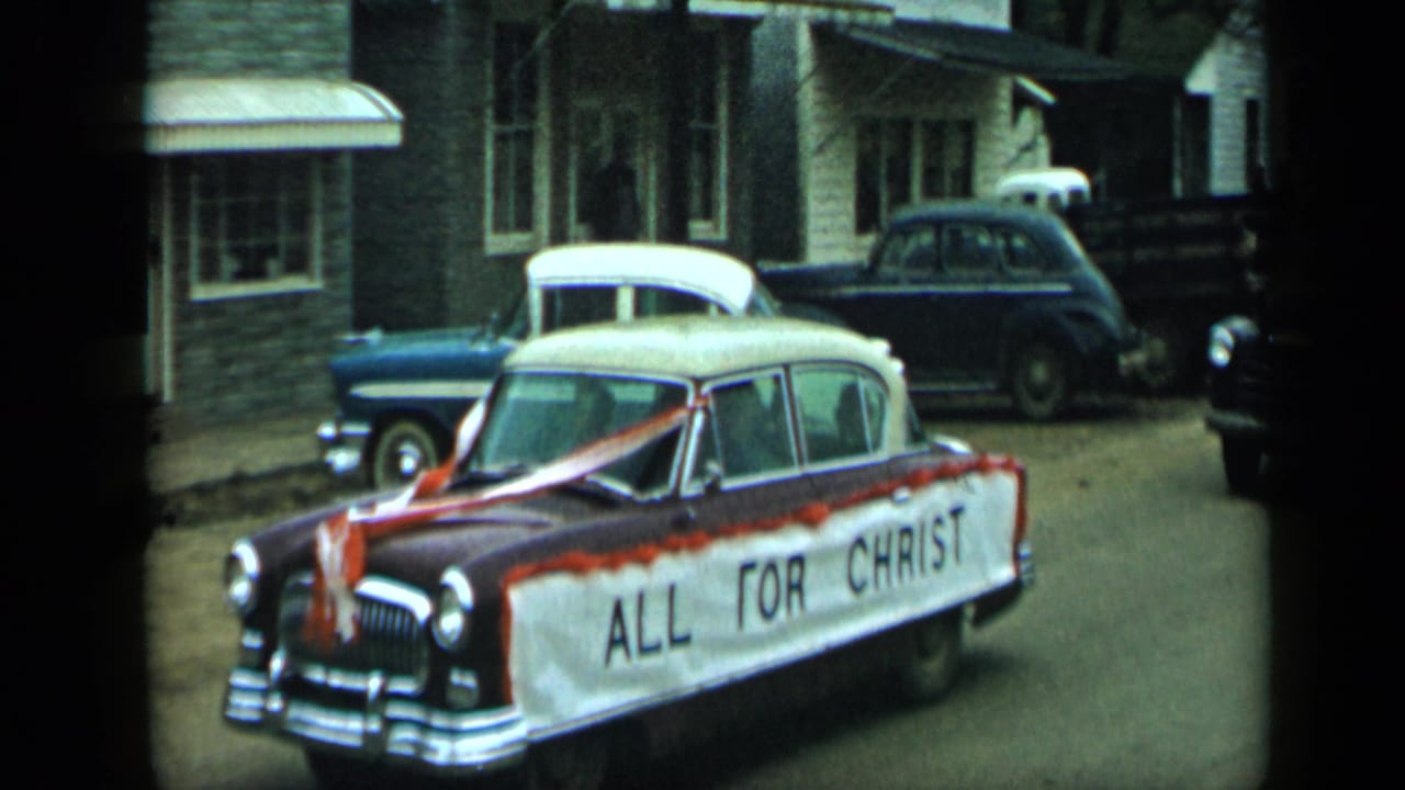 Decorated maroon and white sedan with an ALL FOR CHRIST banner on a small-town street.