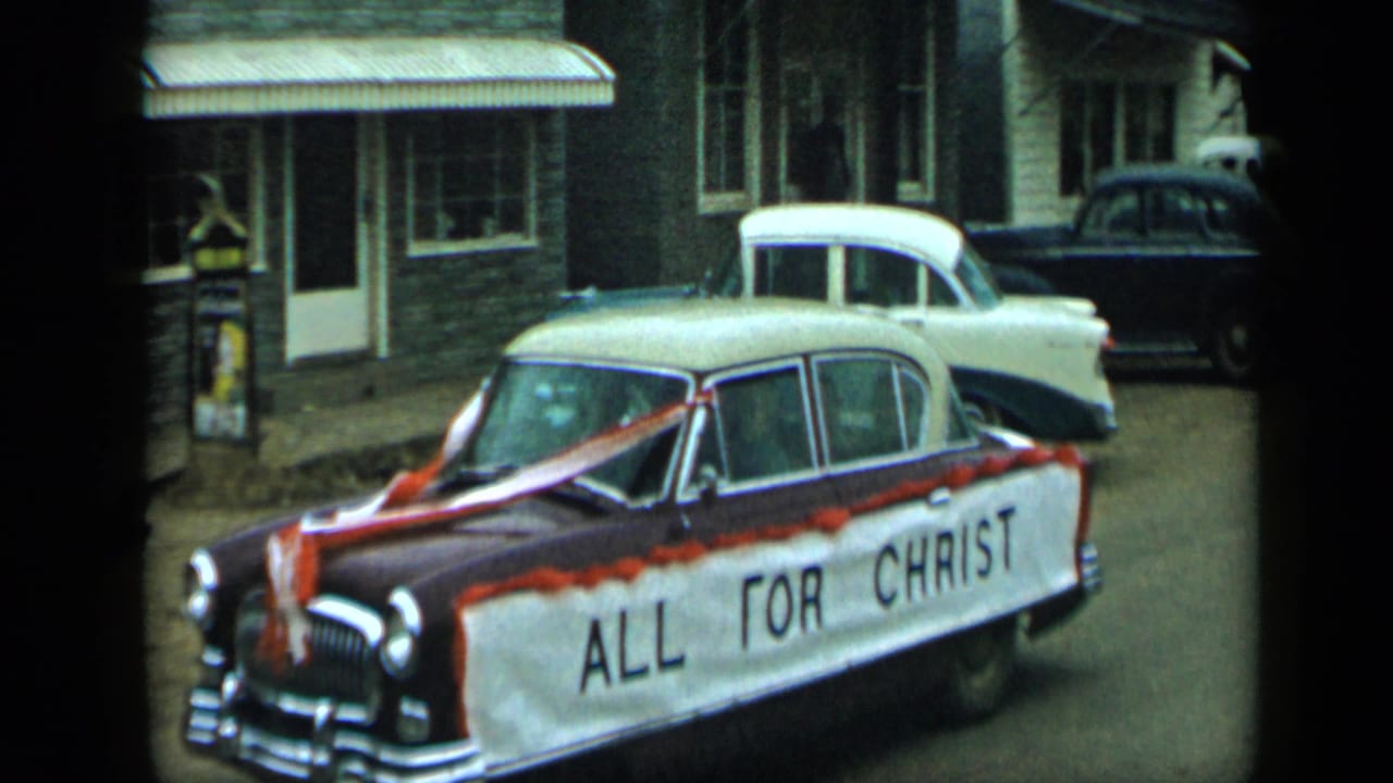 1959 Aledo, Texas parade view of a decorated sedan with an ALL FOR CHRIST banner moving past parked mid-century cars.
